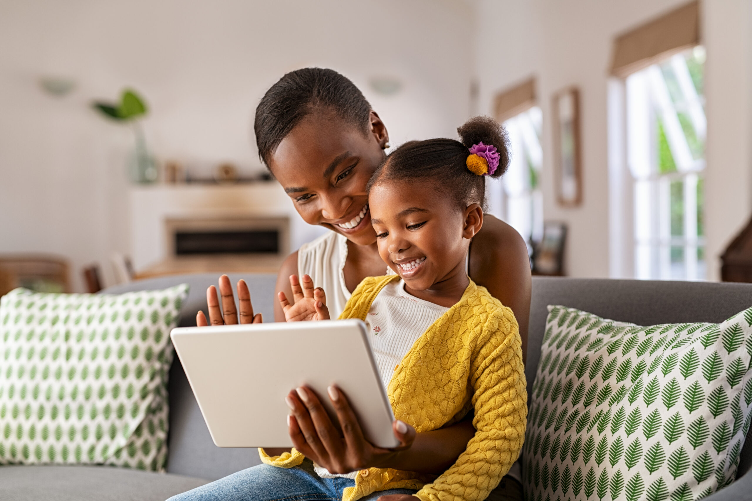 Mother sitting with daughter using a tablet, interacting happily with someone on-screen